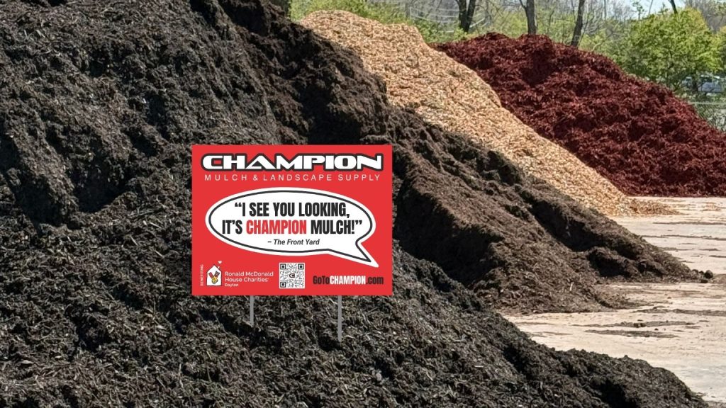 Champion Mulch location with four bulk mulch products lined up in large piles on display and the 2026 Champion Mulch and RMHC Dayton partnership yard sign that has Champion's logo on the top. The words "I see you looking. It's Champion mulch!" in a speech bubble underneath as if the yard is speaking. Under that is the RMHC Dayton logo, a QR code and Champion's web address – go to champion dot com.