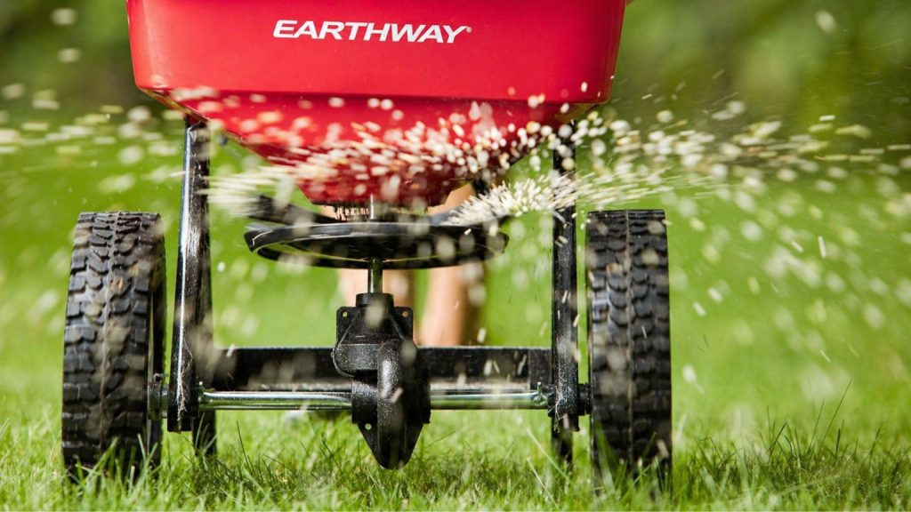  Close-up of a fertilizer spreader moving across a green lawn showing overlapping wheel tracks for even coverage in a Southwest Ohio yard.